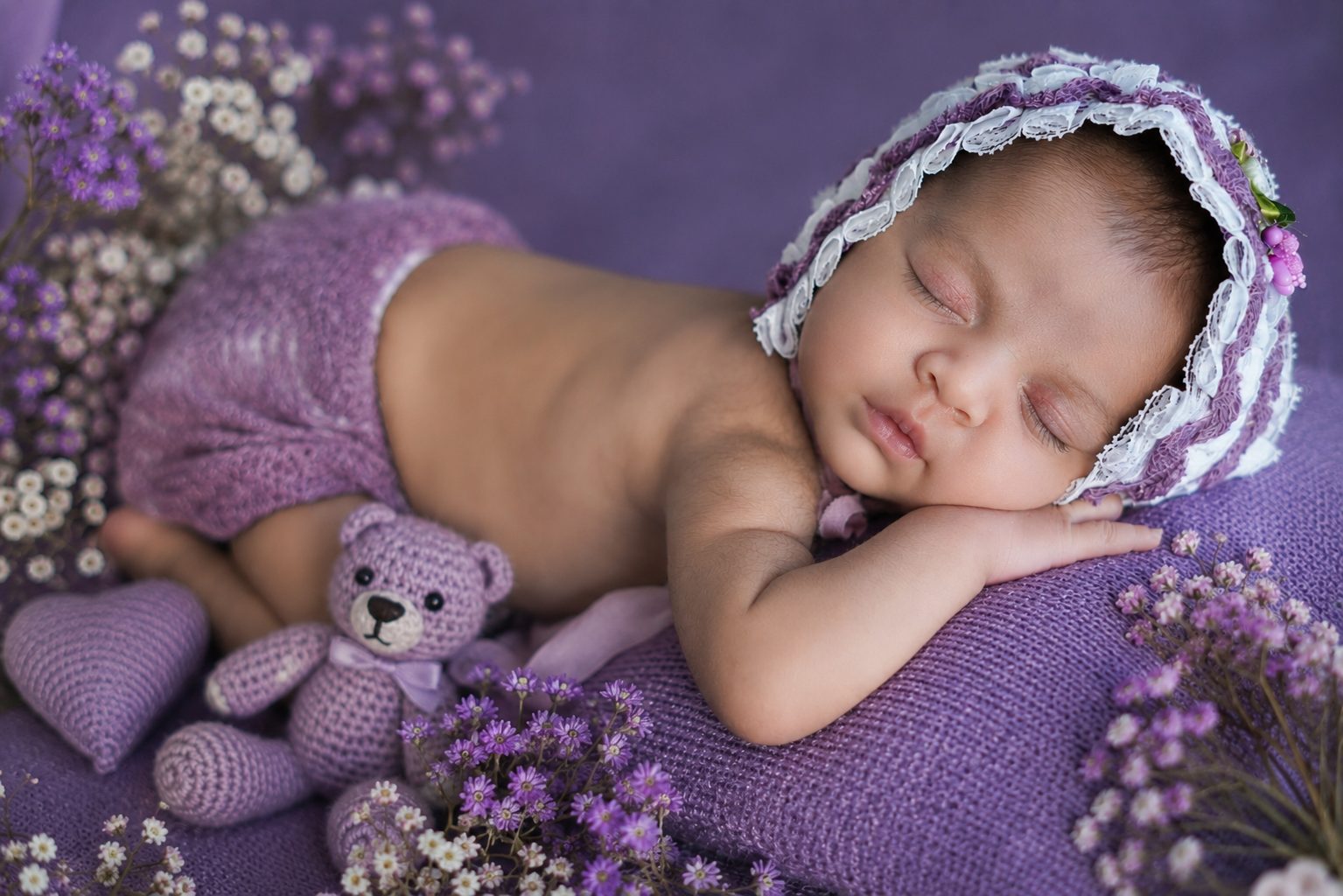 Smiling baby wrapped in a purple blanket beside a crochet teddy bear, surrounded by flowers