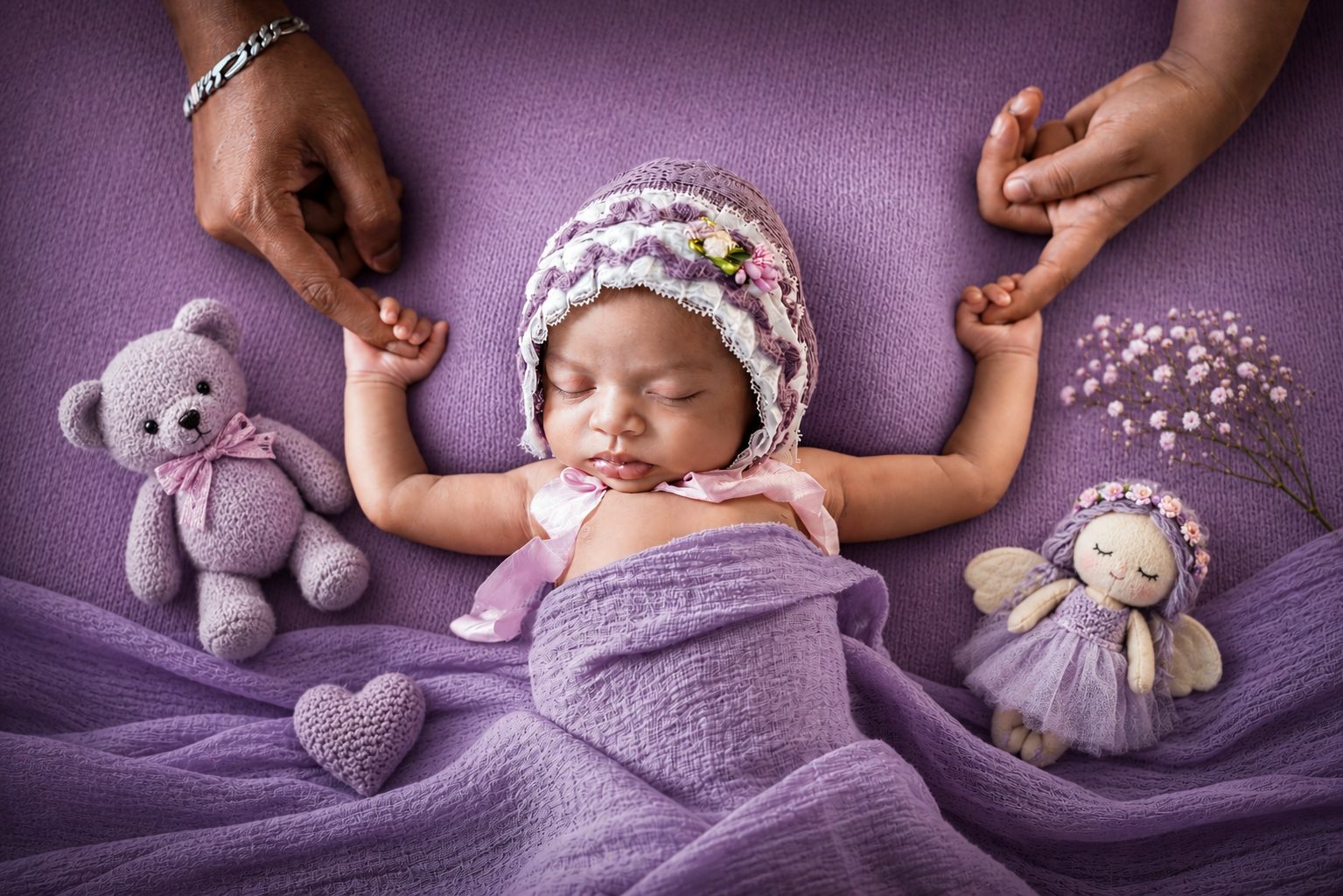 Newborn baby held by parents' hands in a purple-themed setup with teddy bear and flowers