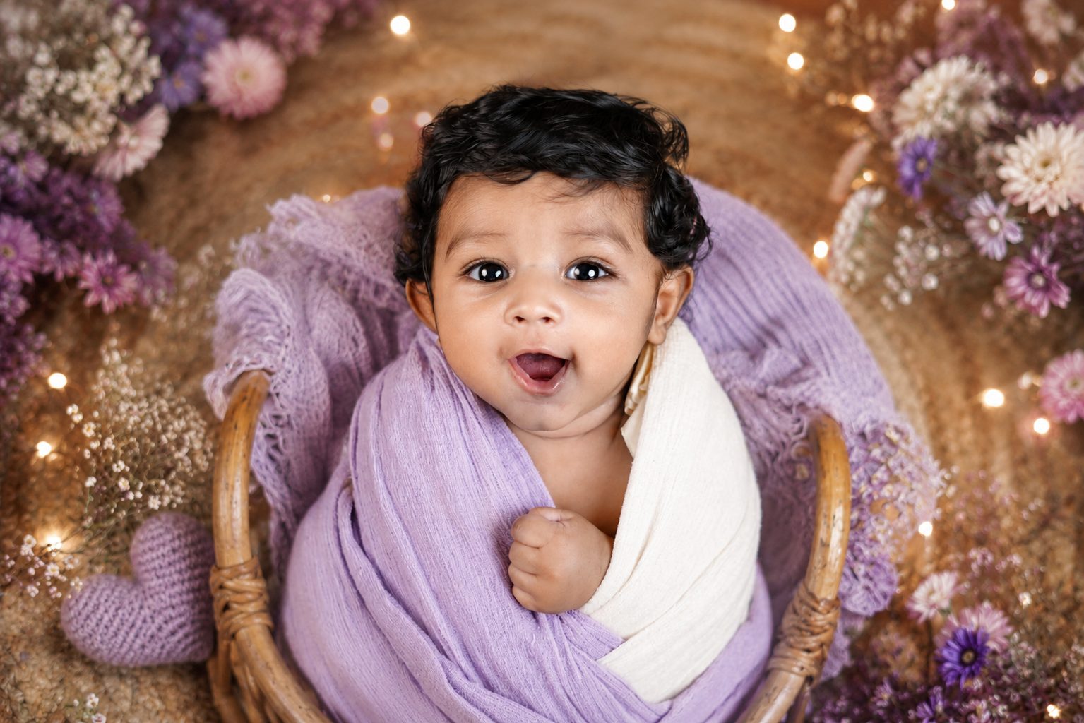 Newborn baby swaddled in a lavender wrap surrounded by pink and lilac roses on cream fur
