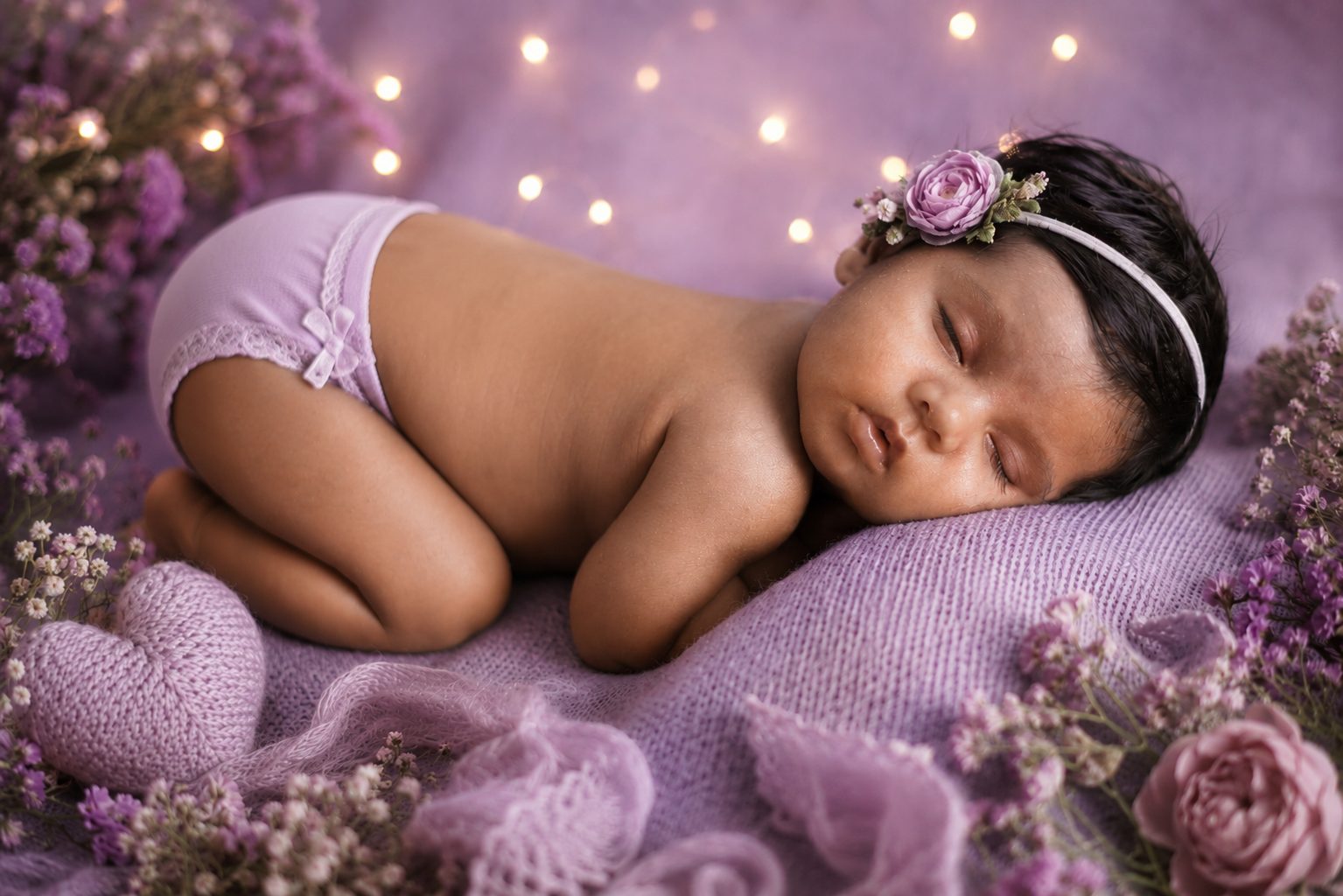 Sleeping newborn in lavender wrap inside a white wicker basket, lit by candlelight and roses